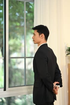 Businessman standing with hands behind back and gazing thoughtfully out of a window.