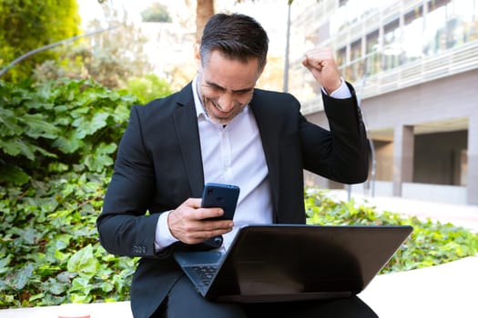 Excited businessman in a suit sitting outdoors, holding a smartphone and raising his fist in celebration. Represents success, achievement, and happiness in professional and digital work.