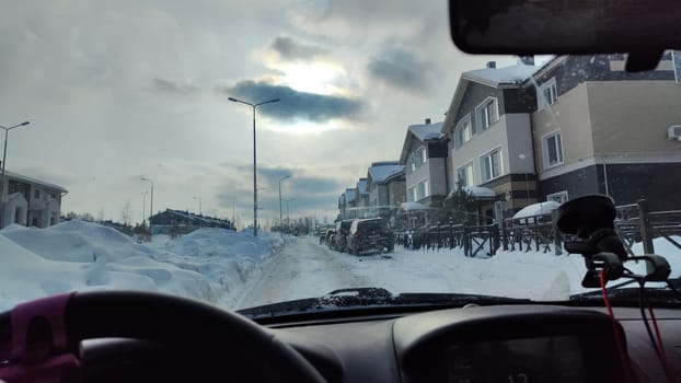 Kirov, Russia - February 2023: Driving through a snow-covered street in a residential area, with houses lining the road and clouds obscuring the sun.