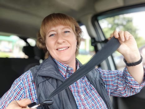 Middle aged woman is seated in a car, smiling cheerfully as she securely fastens her seat belt. Safety in transport. Traffic Laws