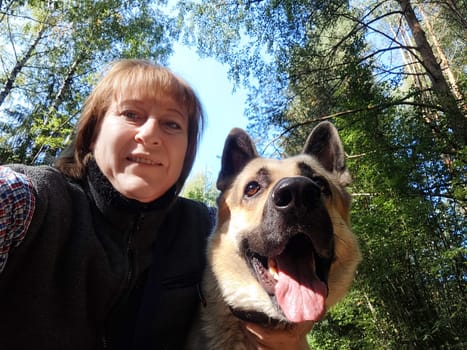 A woman takes a joyful selfie with her friendly German shepherd under the bright sunlight amidst green trees.