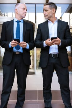 Two middle aged adult men in suits smiling using applications on their mobile phones,conversing outdoors.Vertical