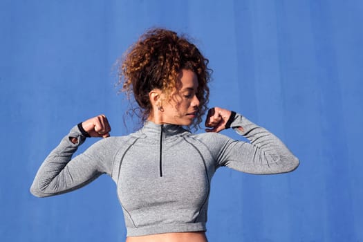 strong female athlete in sportswear with curly hair demonstrating muscles during workout in in front of blue background, concept of sport and healthy lifestyle