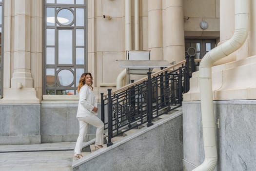 A woman in a white suit walks up a set of stairs outside a building with large windows and a metal railing.