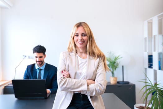 A smiling businesswoman in a suit stands confidently with arms crossed in an office. His colleague works on a laptop in the background, highlighting teamwork and a professional work environment.