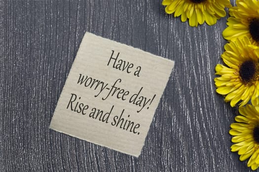 Motivational on brown note with gerbera yellow flower on wooden surface. Have a worry free day, rise and shine.
