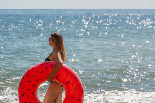 A woman in a bikini holds a large red inflatable ring on a beach with the ocean in the background.