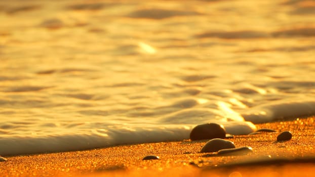 Foamy ocean waves roll onto a sandy beach at sunset, revealing smooth, dark pebbles.