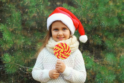 Christmas portrait of happy smiling little girl child in santa red hat with sweet lollipop outdoors on green tree background with snow
