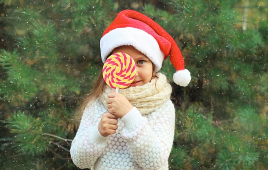 Christmas portrait of happy smiling little girl child in santa red hat with sweet lollipop outdoors on green tree background with snow