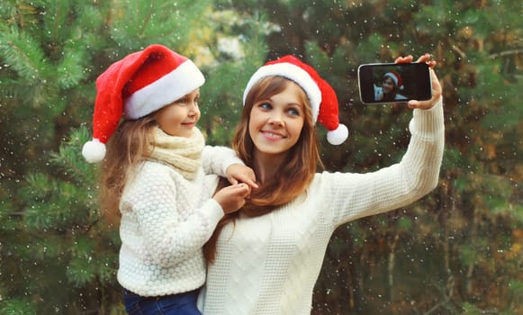 Family, celebration, holiday. Happy mother and little girl child smiling taking selfie with smartphone near Christmas tree in forest together