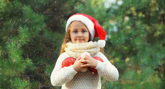 Christmas portrait of happy smiling little girl child in santa red hat with ball toy decoration outdoors on green tree background with snow