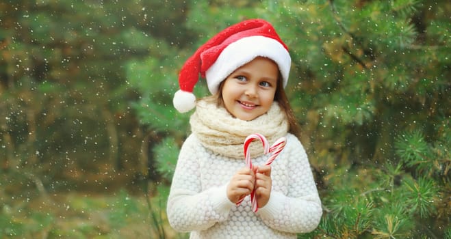 Christmas portrait of happy smiling little girl child in santa red hat with sweet lollipop cane stick outdoors on green tree background with snow
