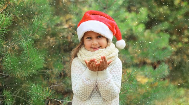 Christmas portrait of happy smiling little girl child in santa red hat blows snow off her hands outdoors on green tree background with snow