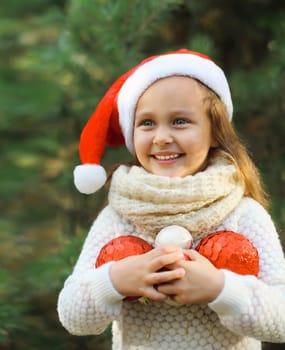 Christmas portrait of happy smiling little girl child in santa red hat with ball toy decoration outdoors on green tree background with snow