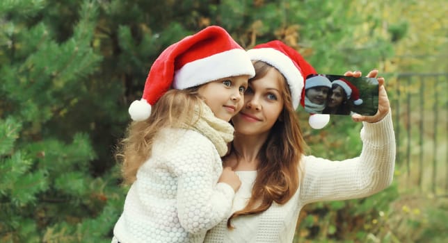 Family, celebration, holiday. Happy mother and little girl child smiling taking selfie with smartphone near Christmas tree in forest together