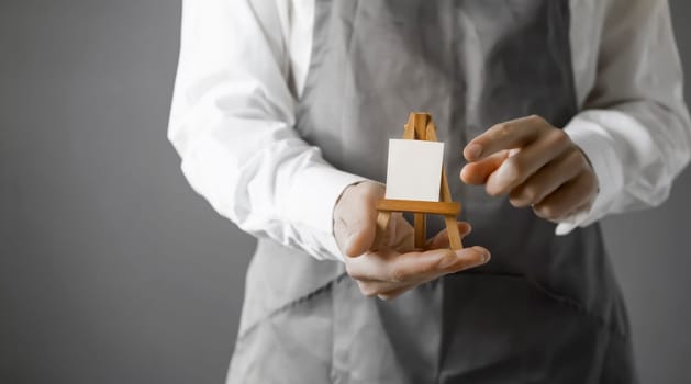 The male artist's hands are holding a miniature easel with a small piece of white paper on it. A creative idea for inviting and discovering art.