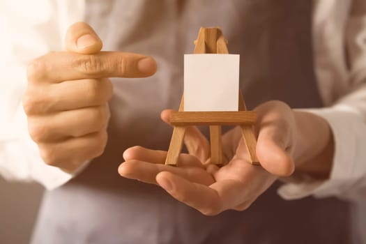 The male artist's hands are holding a miniature easel with a small piece of white paper on it. A creative idea for inviting and discovering art.