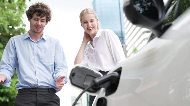 Progressive businessman and businesswoman install charger plug from charging station to electric car before driving around city center. Eco friendly rechargeable car powered by clean energy.