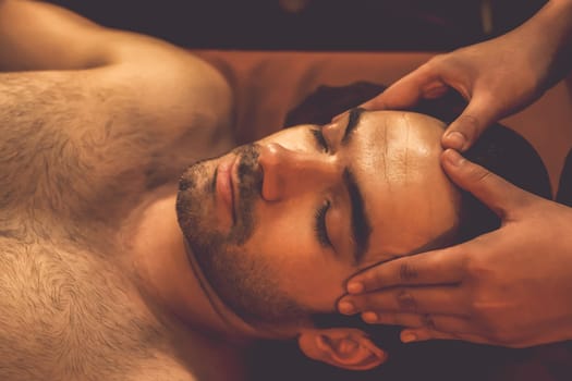 Caucasian man enjoying relaxing anti-stress head massage and pampering facial beauty skin recreation leisure in warm candle lighting ambient salon spa in luxury resort or hotel. Quiescent