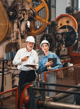 Factory engineer manager with assistant using laptop to conduct inspection of steel industrial machine, exemplifying leadership as machinery engineering inspection supervisor in metalwork manufacture.