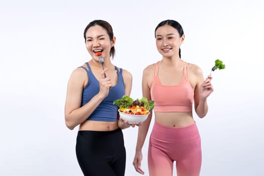 Two young sporty Asian women in sportswear holding salad bowl fill with fruit and vegetable. Natural youthful and fit body lifestyle people with balance nutrition on isolated background. Vigorous