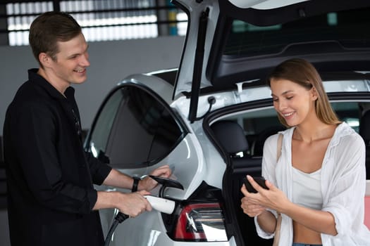 Young couple travel with EV electric car to shopping center parking lot charging in downtown city showing urban sustainability lifestyle by green clean rechargeable energy of electric vehicle innards