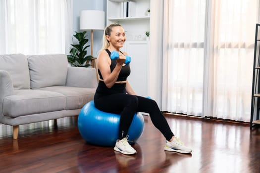 Athletic and sporty senior woman sitting on fit ball while engaging in weight lifting with dumbbell at home exercise as concept of healthy fit body lifestyle after retirement. Clout