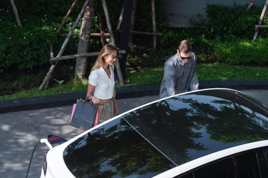 Young couple travel with EV electric car charging in green sustainable city outdoor garden in summer shows urban sustainability lifestyle by green clean rechargeable energy of electric vehicle innards