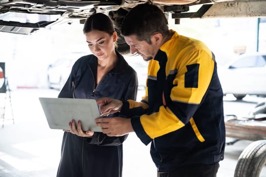 Two vehicle mechanic working together underneath lifted car, conduct car inspection with laptop. Automotive service technician in uniform carefully make diagnostic troubleshooting. Oxus