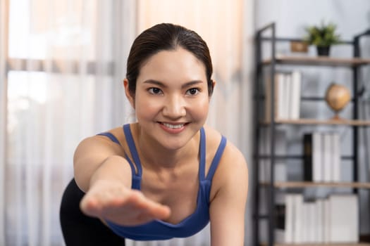 Asian woman in sportswear doing yoga exercise on fitness mat as her home workout training routine. Healthy body care and calm meditation in yoga lifestyle with comfortable and relaxation. Vigorous