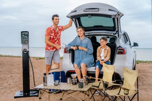 Family vacation trip traveling by the beach with electric car, happy family recharge EV car, enjoying outdoor camping coffee. Seascape travel and eco-friendly car for clean environment. Perpetual