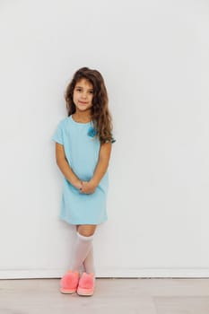 Little Curly Girl In Dress And Pink Slippers Against White Wall