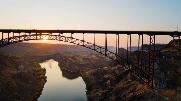 A bridge spans a river with a sunset in the background. The bridge is a metal structure with a wooden deck