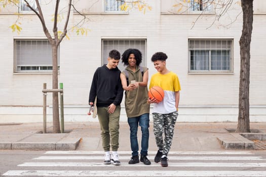 three diverse teen friends with basketball and skateboard talking and smiling happy looking mobile phone while crossing road on crosswalk, concept of youth and urban lifestyle