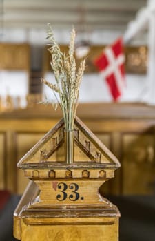 A rustic wooden detail with a vase of wheat indoors. The Danish flag and church setting suggest cultural and historical themes