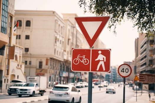 A bustling city street with a bicycle and pedestrian sign, surrounded by buildings and trees. Vehicles and pedestrians share the road in this urban setting