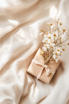 Small gift box decorated with white flowers resting on beige silky fabric, illuminated by natural light