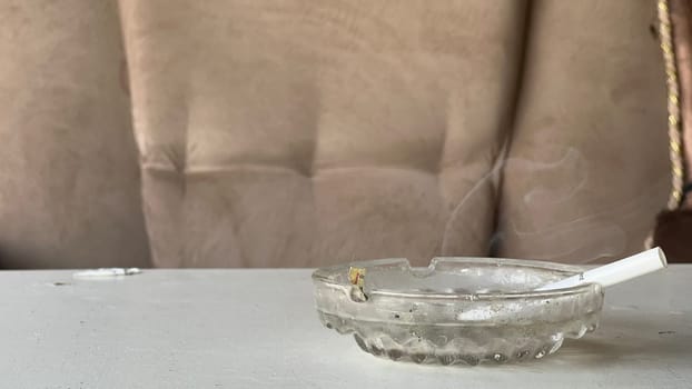 A cigarette rests in an ashtray, producing thin wisps of smoke, highlighting a cozy setting.
