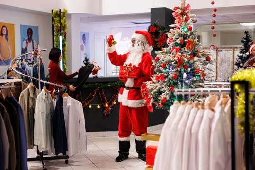 African american man dressed as Santa Claus in festive decorated shopping mall clothing store. Cheerful employee jingling bells and holding red package in fashion boutique shop.