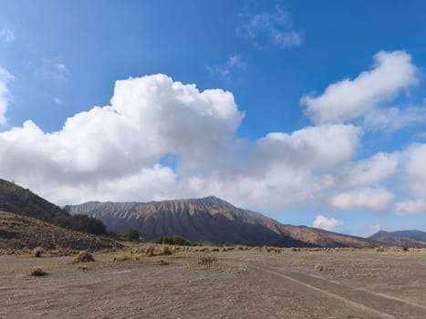 A majestic view of the Bromo Tengger Semeru National Park. The iconic Mount Bromo stands tall with its distinctive crater, surrounded by a vast expanse of volcanic sand and a clear blue sky dotted with fluffy clouds.