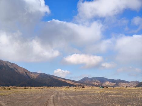 A majestic view of the Bromo Tengger Semeru National Park. The iconic Mount Bromo stands tall with its distinctive crater, surrounded by a vast expanse of volcanic sand and a clear blue sky dotted with fluffy clouds.