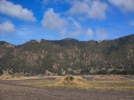 A majestic view of the Bromo Tengger Semeru National Park. The iconic Mount Bromo stands tall with its distinctive crater, surrounded by a vast expanse of volcanic sand and a clear blue sky dotted with fluffy clouds.