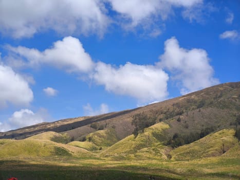 A majestic view of the Bromo Tengger Semeru National Park. The iconic Mount Bromo stands tall with its distinctive crater, surrounded by a vast expanse of volcanic sand and a clear blue sky dotted with fluffy clouds.