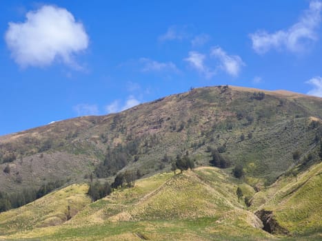 A majestic view of the Bromo Tengger Semeru National Park. The iconic Mount Bromo stands tall with its distinctive crater, surrounded by a vast expanse of volcanic sand and a clear blue sky dotted with fluffy clouds.