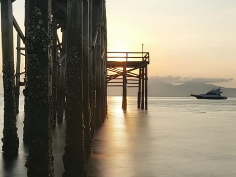 A weathered wooden pier extends into the water at sunrise, with barnacles clinging to the support posts near Banyuwangi, Indonesia