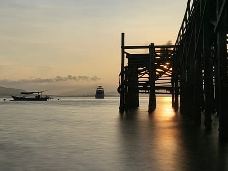A weathered wooden pier extends into the water at sunrise, with barnacles clinging to the support posts near Banyuwangi, Indonesia
