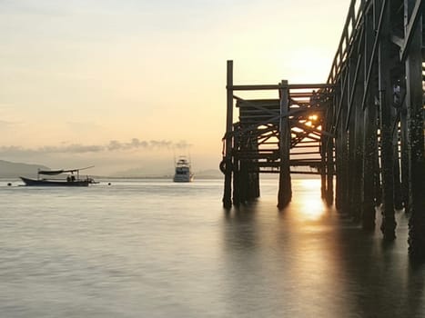 A weathered wooden pier extends into the water at sunrise, with barnacles clinging to the support posts near Banyuwangi, Indonesia