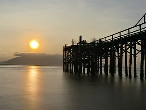 A weathered wooden pier extends into the water at sunrise, with barnacles clinging to the support posts near Banyuwangi, Indonesia