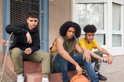 three confident multiracial male teenagers with basketball and skateboard looking at camera sitting together on street, concept of youth and urban lifestyle